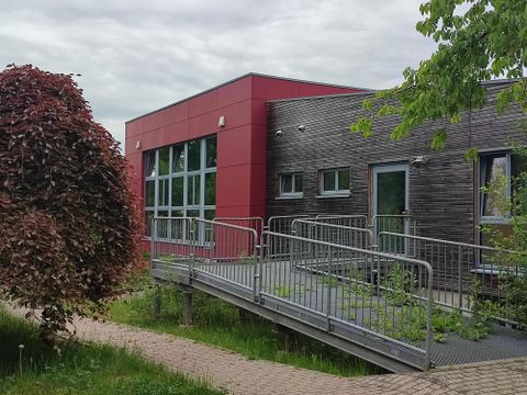 Ein modernes Gebäude mit roter und holzvertäfelter Fassade. Im Vordergrund ist eine Rampenlösung für barrierefreien Zugang zu sehen.