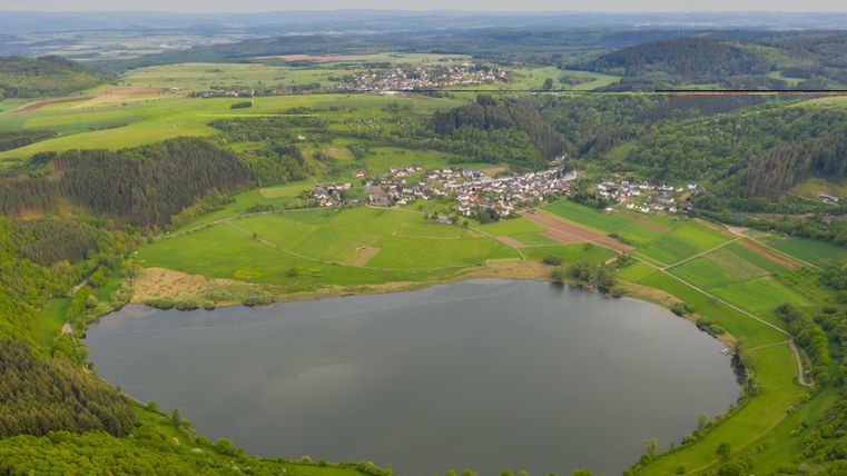 Eine malerische Landschaft mit einem ruhigen See und üppigem Grün. Im Hintergrund erheben sich sanfte Hügel und ein kleines Dorf ist sichtbar.