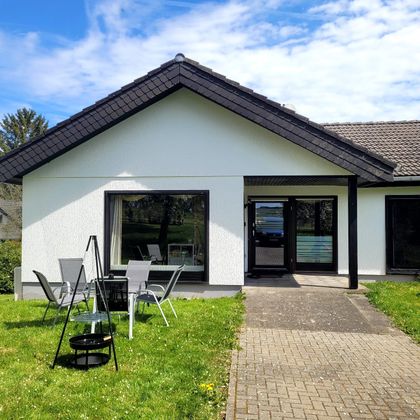 Ein modernes Einfamilienhaus mit einer hellen Fassade und einem geschmackvollen Vorgarten. Vor der Tür befinden sich Gartenmöbel und ein Grill.