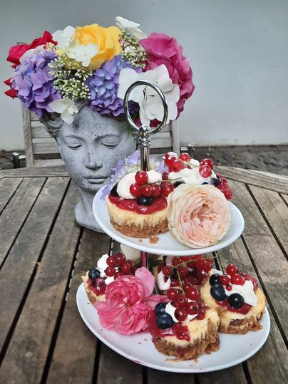 Ein Tisch mit einer Etagere voller kleiner Kuchen, dekoriert mit Beeren und Blumen. Im Hintergrund eine Blumenvase in Form eines Kopfes.