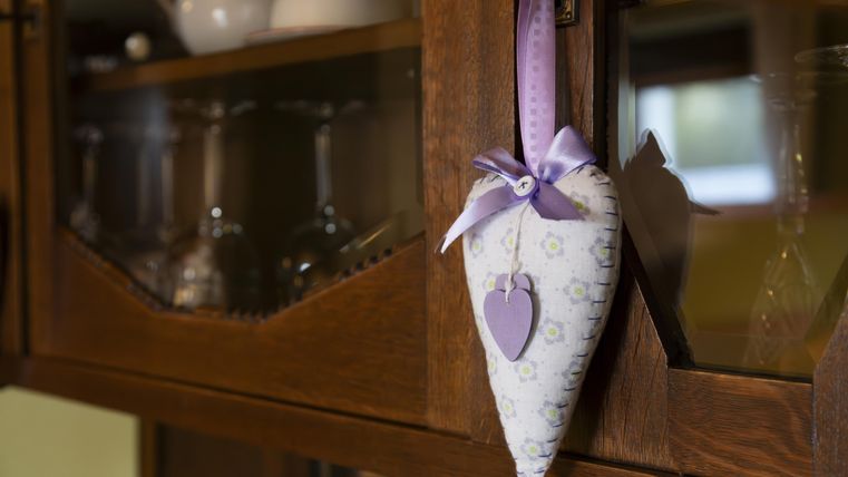 Een decoratief stoffen hart hangt aan een keukenkast. Het is versierd met een paarse strik en een kleine paarse hartbedel.