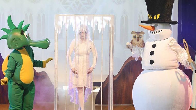 Une performance théâtrale colorée avec un dragon, un bonhomme de neige et une femme captive dans une cage glacée. Les personnages portent des costumes frappants et se tiennent sur une scène festive.