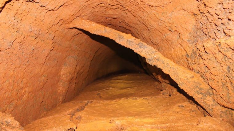 Een smalle tunnel met roodbruine wanden en een ruw oppervlak. De vloer bestaat uit een mineraal materiaal dat tussen de wanden lijkt te zijn ingeklemd.