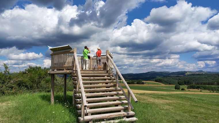 Houten platform met twee mensen die naar het landschap kijken onder een bewolkte hemel.