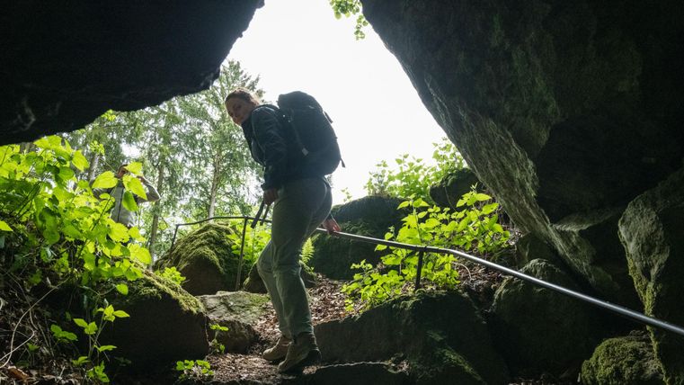 Two people are climbing out of a cave into the open air. Surrounded by rocks and plants, a railing leads the way up.