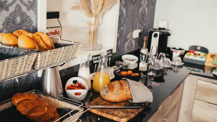 Une table de petit-déjeuner soigneusement dressée avec du pain frais, des croissants et diverses boissons. En arrière-plan, des éléments de décoration et un aménagement de cuisine moderne sont visibles.