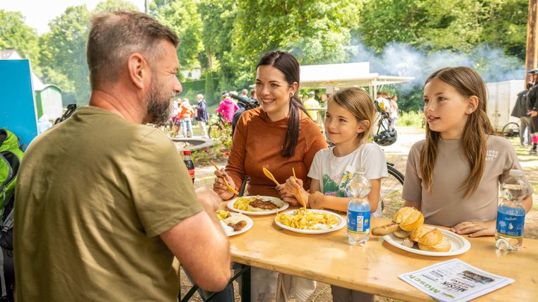 Een blije familie zit aan een tafel buiten en geniet samen van hun maaltijd. Twee kinderen en twee volwassenen lachen en praten met elkaar bij mooi weer.