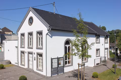 Un bâtiment moderne, blanc, avec de grandes fenêtres et un toit en pente. L'extérieur est bien entretenu avec de petits arbres et un chemin.