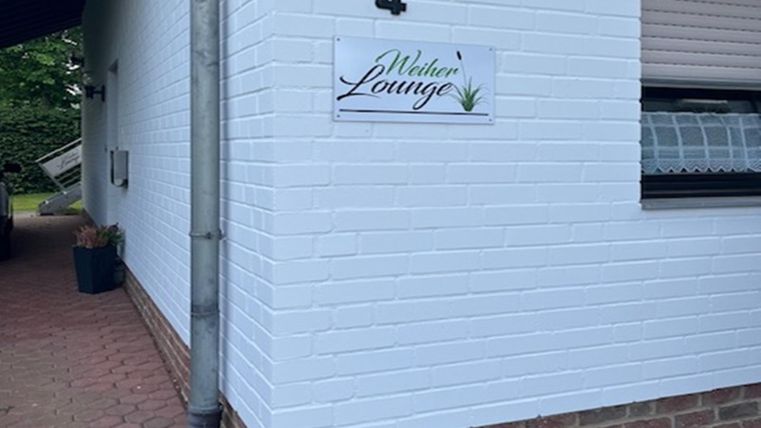 Une maison blanche avec un mur clair et une petite enseigne portant l'inscription « Weberei Lounge ». À côté de l'entrée se trouve un pot de fleurs vert.