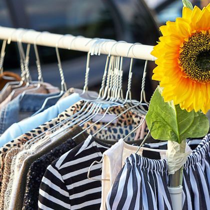 Een kledingrek met verschillende kledingstukken, waaronder gestreepte tops en broeken. Naast de kleding staat een zonnebloem, die het beeld opfleurt.