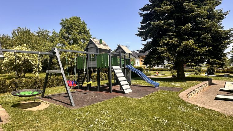 A playground with a slide, a swing, and a climbing frame.
Surrounded by green grass and trees under a shining blue sky.