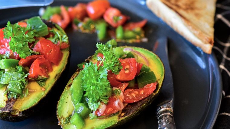 Filled avocado halves with tomatoes and peppers on a plate, next to a piece of toast.