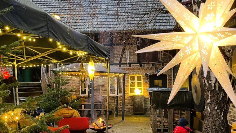 Een gezellige tuinlandschap met lichtjes, een kerstster en een kampvuur. Op de achtergrond zijn historische gebouwen en kerstdecoratie te zien.