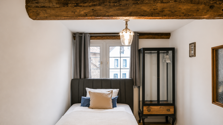 Une chambre confortable avec un lit simple et une étagère en bois. La fenêtre offre une vue sur une rue calme et la pièce dégage une atmosphère chaleureuse et accueillante.