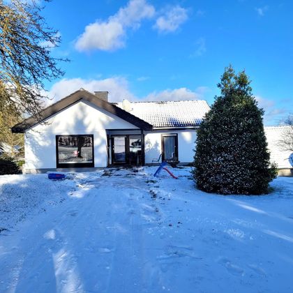 Een modern huis in de sneeuw met een groene haag en een slee op de voorgrond. De lucht is blauw met enkele wolken.