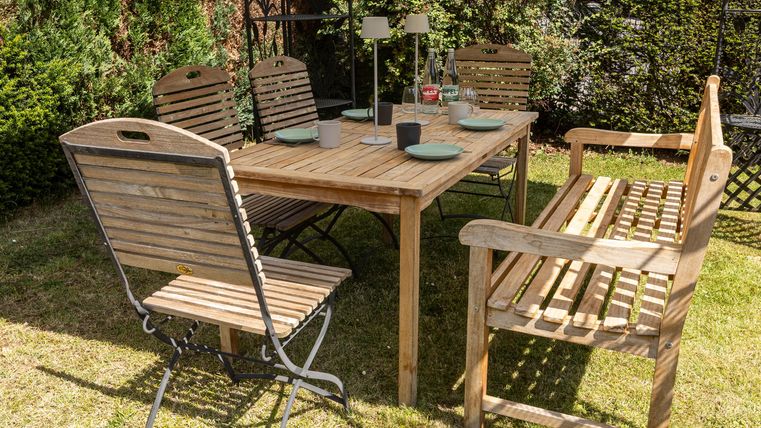 Een houten tafel met stoelen staat in een tuin. De tafel is voorbereid voor een maaltijd buiten.