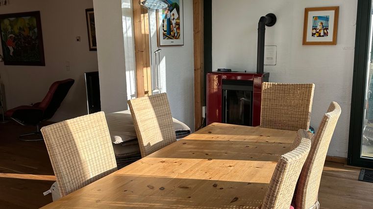 A bright dining room with a large wooden table and several chairs. In the background, cozy furniture and a fireplace are visible.
