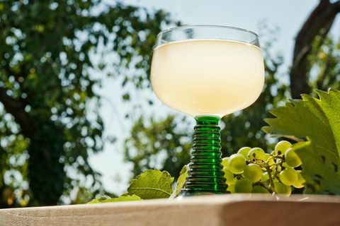 An elegant glass with a clear drink is on a table. In the background, green leaves and grapes can be seen.