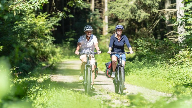 Zwei Radfahrerinnen fahren auf einem schmalen Weg durch einen grünen Wald. Die Sonne scheint und es ist eine freundliche, entspannte Atmosphäre.