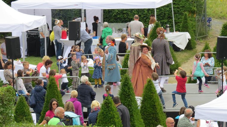 Une fête animée avec divers stands et des gens qui dansent et discutent. Certaines personnes portent des costumes historiques, tandis que des enfants et des adultes s'amusent ensemble.