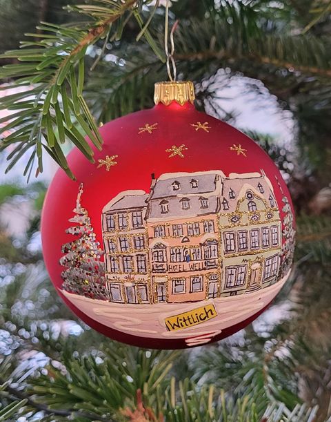 Eine rote Weihnachtskugel mit einer Darstellung von Häusern und einem Weihnachtsbaum. Sie zeigt die Stadt Wittlich und ist mit goldenen Sternen verziert.