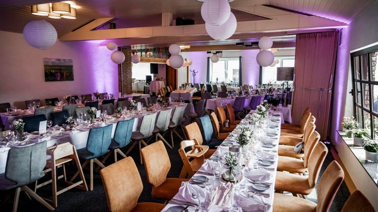 An elegantly decorated hall with festively set tables. Soft purple lighting creates an inviting atmosphere for events.