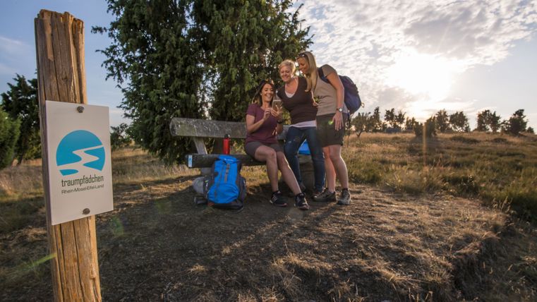 Drei Frauen sitzen auf einer Bank in einer Heidelandschaft mit einem Schild "traumpfädchen" im Vordergrund.