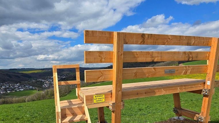 Un banc en bois avec des marches se trouve sur une colline surplombant un paysage. Le ciel est partiellement nuageux et le vert des prairies est bien visible.