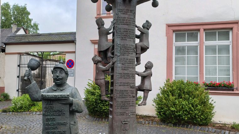 Een sculptuur van een boom met kinderen die erin zitten, en een figuur die een bord vasthoudt. Op de voorgrond spuiten kleine waterfonteinen en groene planten omringen het.