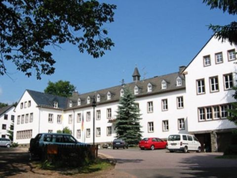 Uitzicht op een groot drieverdiengebouw in L-vorm. Op het zwart gedekte dak zijn elf dakkapellen geplaatst. Achter het gebouw steekt de top van een klokkentoren uit.
Voor het gebouw is er een parkeerplaats met meerdere auto's.