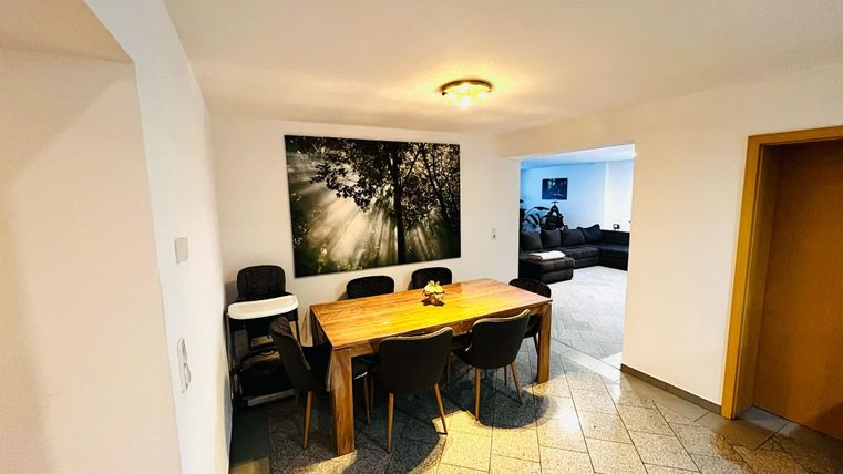 Een moderne eetruimte met een houten tafel en zwarte stoelen. Aan de muur hangt een afbeelding met zonnestralen door bomen.