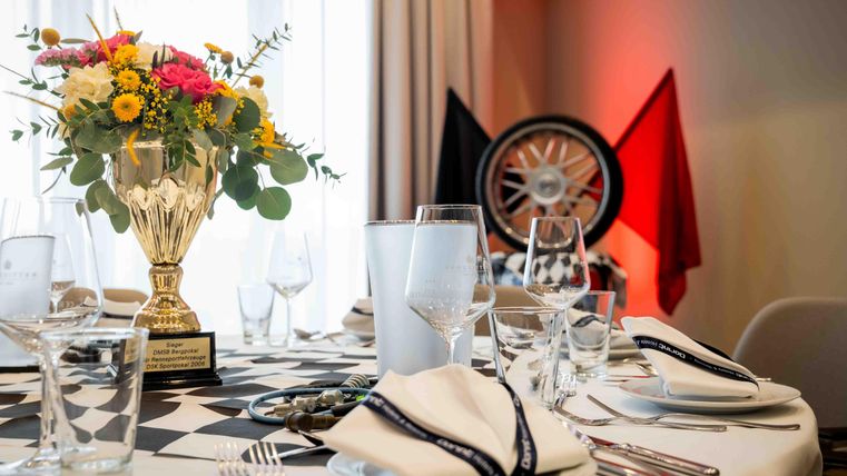 A festively set table with a floral arrangement in a trophy and glasses. In the background are black and red flags as well as a wheel.