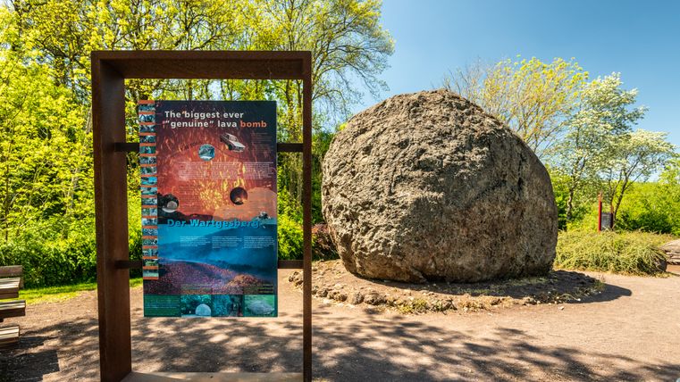 Eine große Lavabombe in Strohn, umgeben von Bäumen und einem Informationsschild.