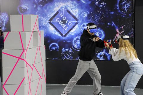 Twee personen spelen een virtual-reality-spel in een moderne omgeving. Op de achtergrond zijn opvallende graphics en een structuur van kartonnen dozen met roze markeringen te zien.