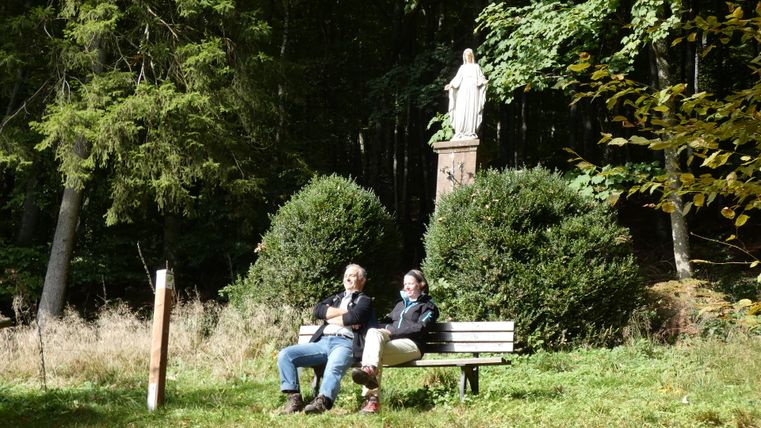Deux personnes sont assises sur un banc devant une colonne mariale dans la forêt.