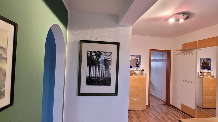 Ein heller Flur mit Holzfußboden und grüner Wand. An der Wand hängen Bilder, und es gibt Schränke und Türen im Hintergrund.