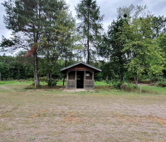 Small wooden hut in the forest, surrounded by trees and a grassy area. The sky is cloudy., © TI Bitburger Land - Steffi Wagner