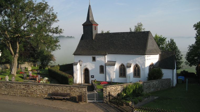 White chapel with pointed tower, surrounded by cemetery and trees, in the morning mist.