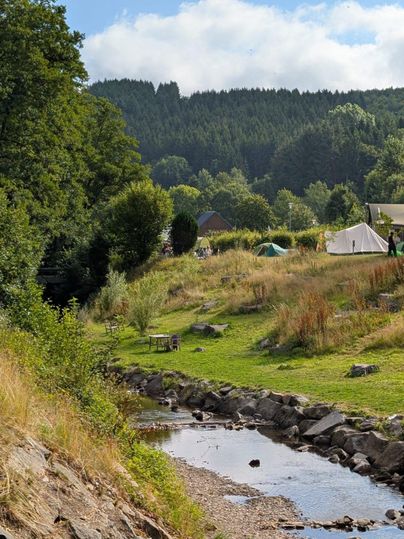 Un paysage paisible avec une petite rivière et des collines douces. Sous les arbres, on peut voir des tentes et des sièges.