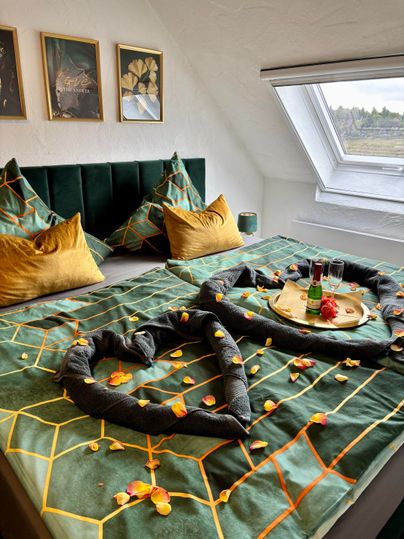 An elegant bedroom with a large bed, decorated with green bedding and golden pillows. On the bed lies a romantic arrangement of towels and flower petals, next to it stands a tray with drinks and snacks.
