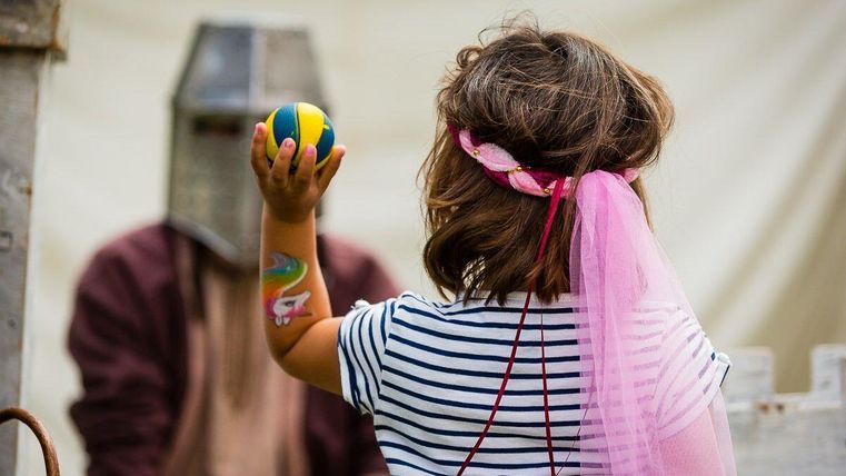 Ein Kind trägt ein buntes Tuch und hält einen Ball in der Hand. Im Hintergrund ist eine Person in einer Rüstung zu sehen.