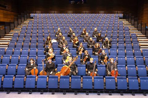 An orchestra is sitting on blue chairs in a hall. The musicians are holding their instruments and are ready to play.