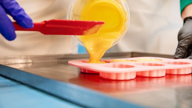 A person is pouring liquid yellow mass into silicone molds. The hands are protected with gloves and a colored apron.