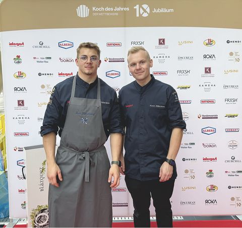 Twee mannen in kookkleding en schort staan glimlachend voor een sponsorswand