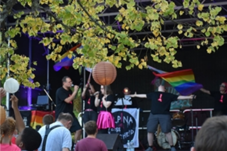 Een levendig podium met artiesten die voor een publiek zingen. Regenboogvlaggen wapperen en er heerst een feestelijke sfeer.
