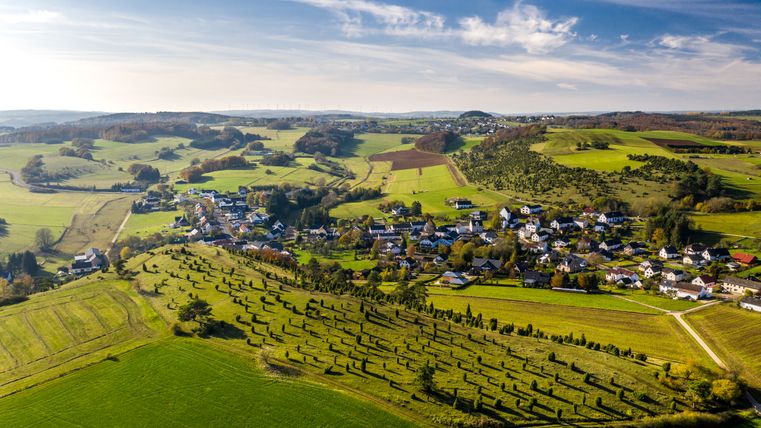 Luftaufnahme einer grünen Hügellandschaft mit einem Dorf in der Eifel.