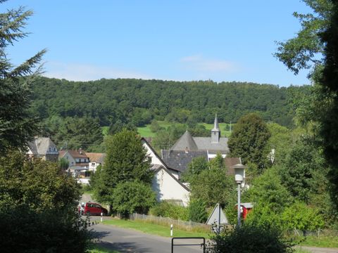 Ein malerisches Dorf umgeben von üppigem Grün und sanften Hügeln. Im Hintergrund ist eine Kirche mit einem hohen Turm zu sehen.