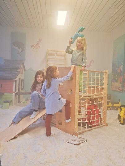 Een speelgebied met drie meisjes die op een speeltoestel spelen. De grond is bedekt met zand en er zijn speelgoed en een kleine glijbaan te zien.