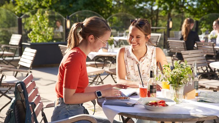 Twee vrouwen zitten vrolijk aan een tafel in de open lucht en genieten van hun tijd samen. Op de tafel staan drankjes en een klein bord met eten.