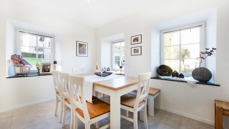 Ein heller Essbereich mit einem weißen Tisch und Stühlen. Große Fenster sorgen für viel Tageslicht und eine gemütliche Atmosphäre.